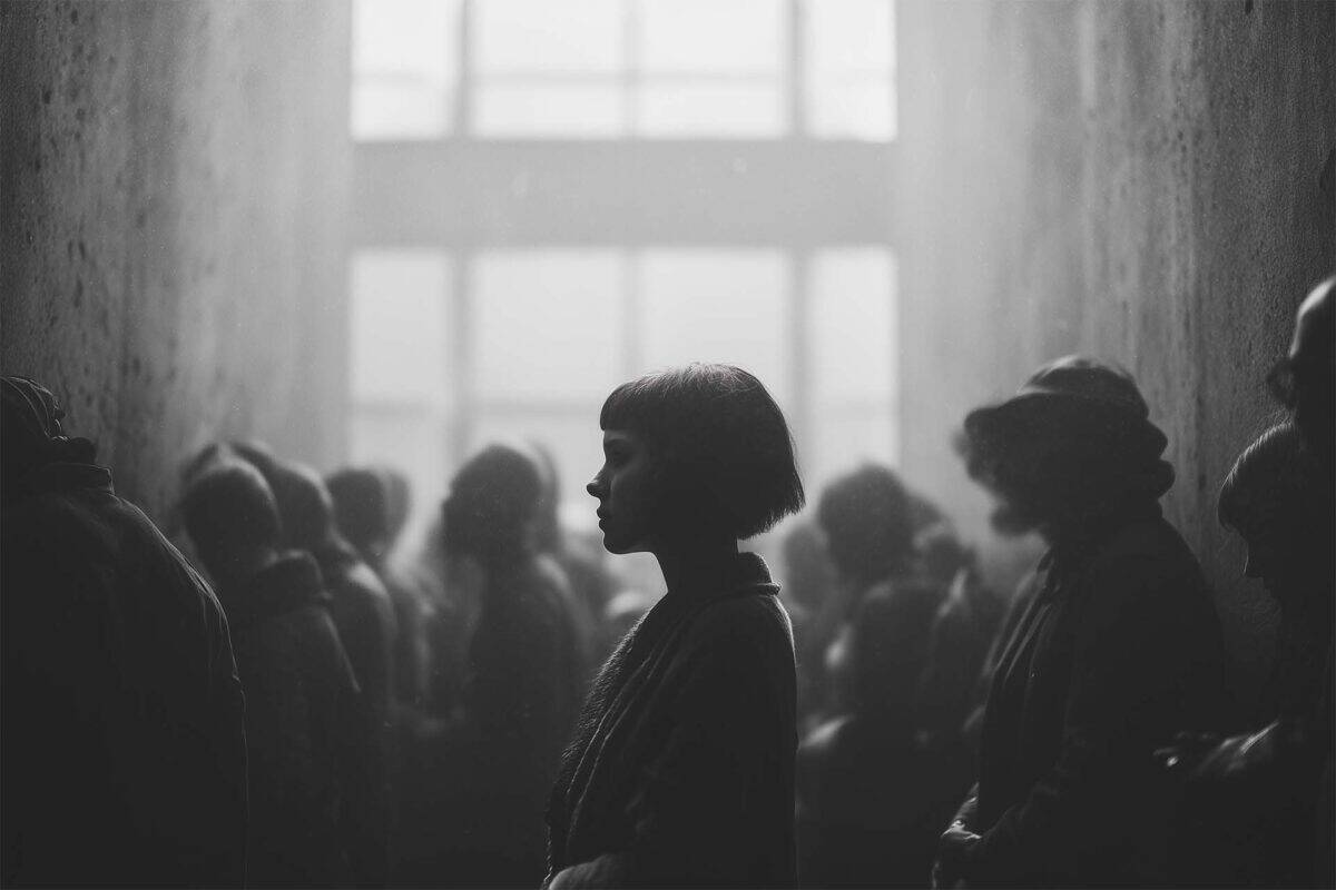 You could arrive hours early and still be behind in line. Woman stands still in profile, centered and surrounded by a multi-row crowd in soft blur against brutalist concrete walls.